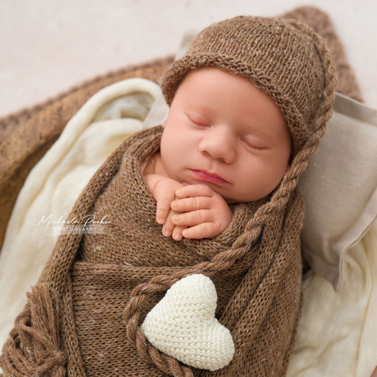 Schlafendes Neugeborenes, eingewickelt in braune Stricktücher mit passender Haube, hält die Hände gefaltet und liegt neben einem kleinen gehäkelten Herz.